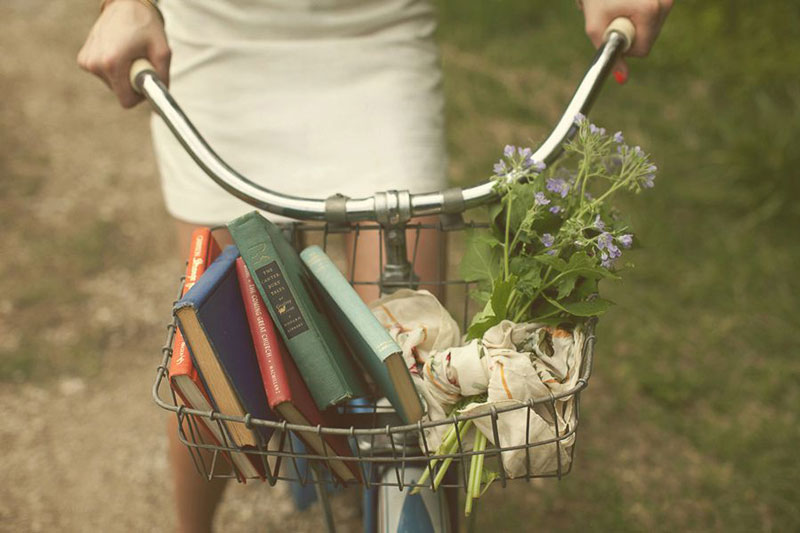 κοπέλα πάνω σε ποδήλατο με καλαθάκι που έχει μέσα λουλούδια και βιβλία