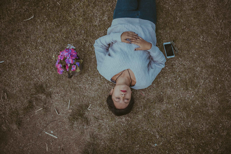 γυναίκα συλλογιέται τον φόβο του θανάτου ξαπλωμένη στο χώμα έχει δίπλα της λουλούδια και ένα κινητό