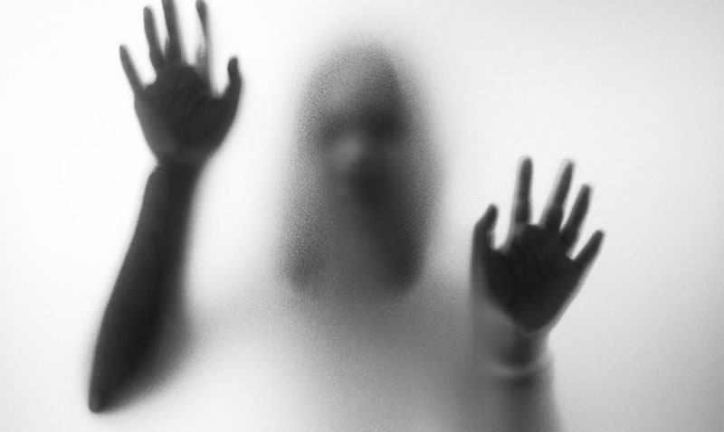 ασπρόμαυρη φωτογραφία γυναίκας που ακουμπά τα χέρια της σε τζάμι που αντιμετωπίζει σιωπηρή κακοποίηση στην σχέση της