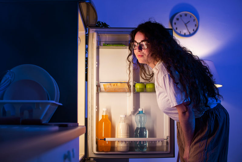γυναίκα στέκεται μπροστά στο ανοιχτό ψυγείο της το βράδυ