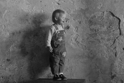 ασπρόμαυρη φωτογραφία που απεικονίζει ένα αγοράκι να στέκεται δίπλα σε έναν φθαρμένο τοίχο