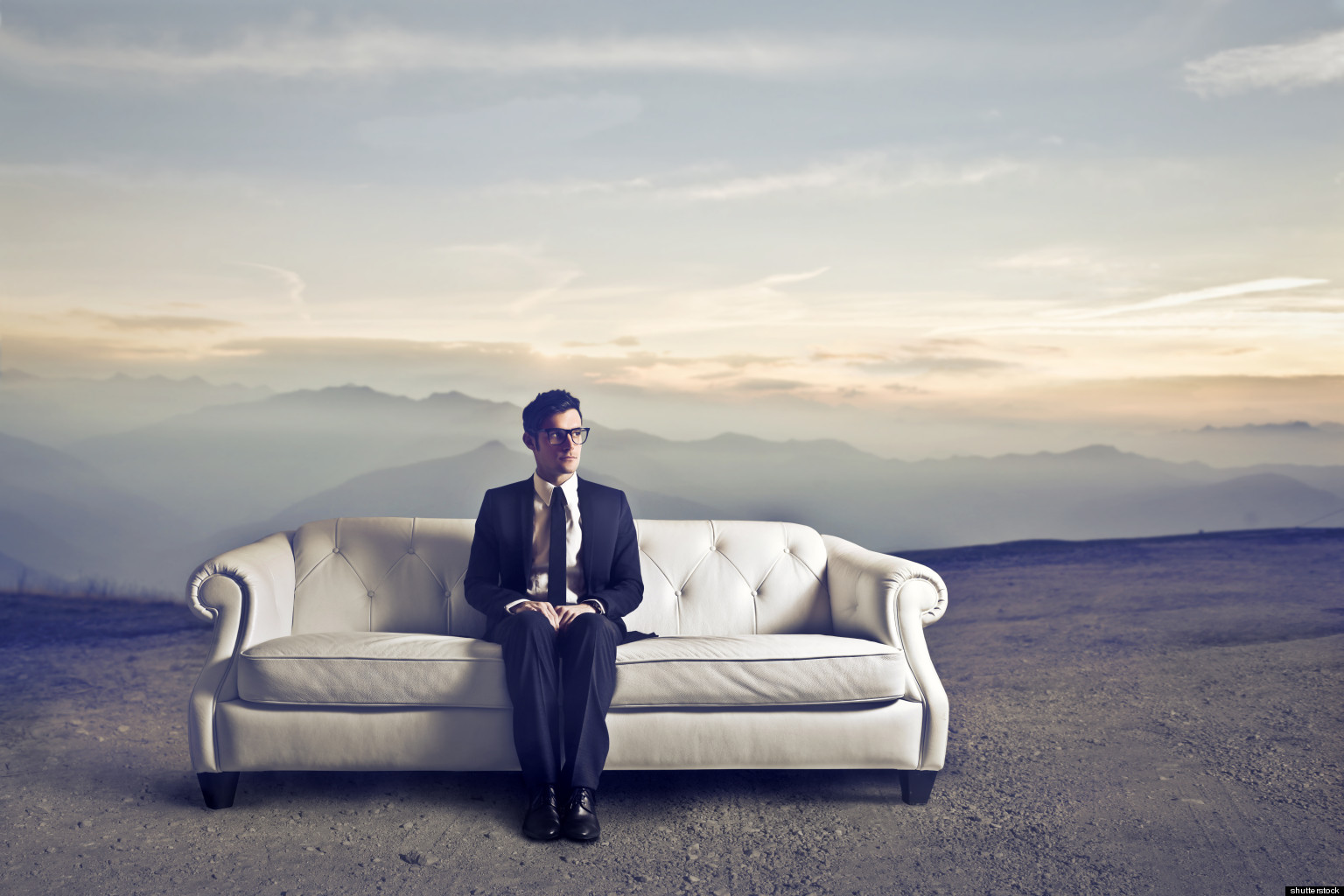 άντρας με κοστούμι και γυαλιά κάθεται μόνος του σε έναν καναπέ που βρίσκεται στην έρημο