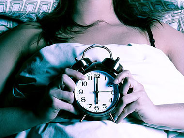 γυναίκα ξαπλωμένη στο κρεββάτι κρατάει ένα ξυπνητήρι