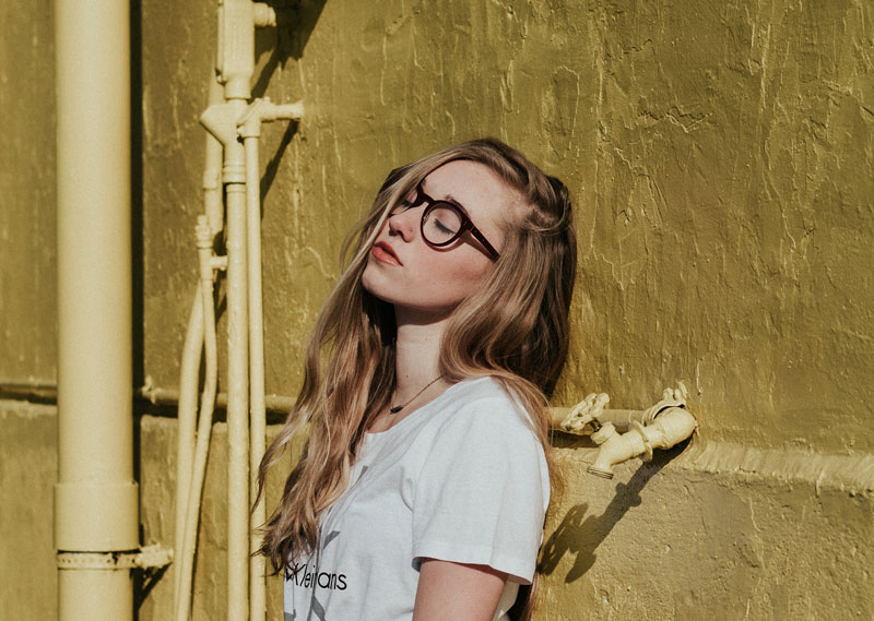 κορίτσι με γυαλιά και λευκό μπλουζάκι που στέκεται με την πλάτη σε ένα τοίχο και έχει κλειστά μάτια